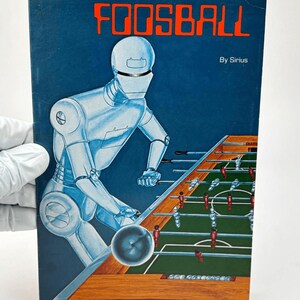 May include: A vintage book cover titled Computer Foosball by Sirius, featuring a blue background with a large silver robot playing a foosball table. The table has a green surface with red and silver player figures and a wooden frame.