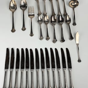 May include: A collection of silver-toned flatware including twelve dinner knives with pointed blades, five forks, five spoons, one small fork, one ladle, and one butter knife. Each piece features a smooth, polished finish and a simple handle design.