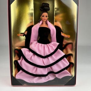 Puede incluir: Una muñeca con el cabello oscuro recogido en un moño, vistiendo un corpiño negro sin tirantes y una falda rosa de varias capas con bordes negros. La muñeca se presenta en una caja rectangular con un fondo dorado y una ventana frontal.