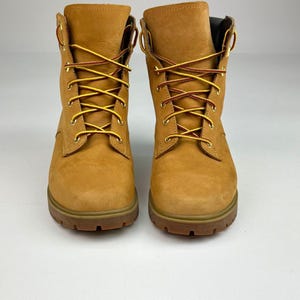 Puede incluir: Un par de botines de cuero color canela con punta redondeada, cierre frontal de cordones con ojales de metal dorado y suelas de goma marrón gruesas y resistentes. Los botines están sujetos con cordones estampados en amarillo y marrón.