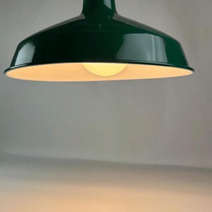May include: A pendant light fixture featuring a wide, dome-shaped shade with a glossy dark green exterior finish and a smooth white interior. A single round, warm-toned light bulb is visible inside the center of the shade, suspended by a cord.