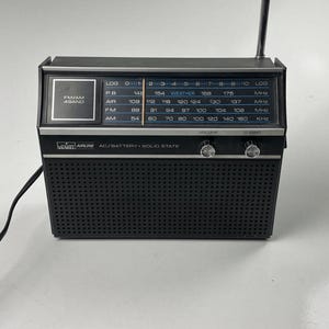 Könnte beinhalten: Ein schwarzes tragbares Radio mit silbernem Rand, das ein Frequenzanzeigefeld mit FM-, AM-, AIR- und PB-Bändern aufweist. Die Vorderseite enthält ein Lautsprechergitter, zwei silberne Drehknöpfe für Lautstärke und Sendersuche sowie eine Antenne.