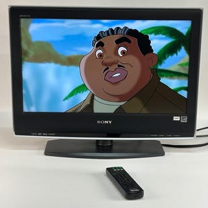 May include: A black Sony Bravia flat-screen television with a silver bottom trim and a black pedestal base. The screen displays a colourful animated character against a tropical background. A black rectangular remote control sits in front of the television.