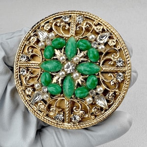 May include: A round gold-tone brooch featuring a central cluster of marquise-cut green stones and small white pearls. The design includes intricate filigree metalwork, clear faceted crystals, and a textured border, creating a detailed accessory.