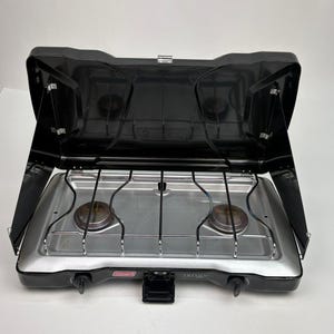 Può includere: Un fornello da campeggio portatile Coleman Triton a due fuochi, nero e argento, con coperchio incernierato, paravento laterali, superficie di cottura argentata, due bruciatori circolari, una griglia cromata e due manopole nere sul pannello frontale.