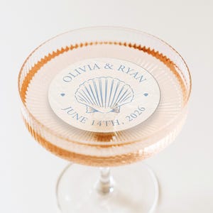 May include: A champagne coupe glass filled with a pink beverage, with a circular coaster featuring a seashell design and the names "Olivia & Ryan" and the date "June 14th, 2026". The glass has a ribbed design.