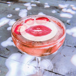 May include: A pink cocktail with a lipstick kiss printed on the top. The cocktail is garnished with strawberry slices.