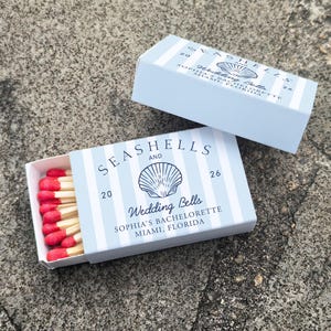 May include: Two light blue matchboxes with white vertical stripes and the words "Seashells and Wedding Bells" printed on them. The matchboxes contain red-tipped matches. The text "Sophia's Bachelorette, Miami, Florida" is also printed on the boxes.