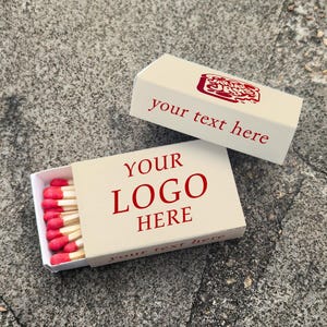 May include: Two beige matchboxes with red text and matches. One box is open, revealing red-tipped matches. The text on the open box reads "YOUR LOGO HERE". The other box has the text "your text here".