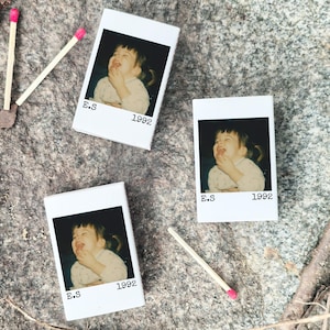 May include: Three white matchbox-style containers with a photo of a laughing child, the text "E.S 1992". Two matches with pink tips are next to the containers. The items are on a grey stone surface.