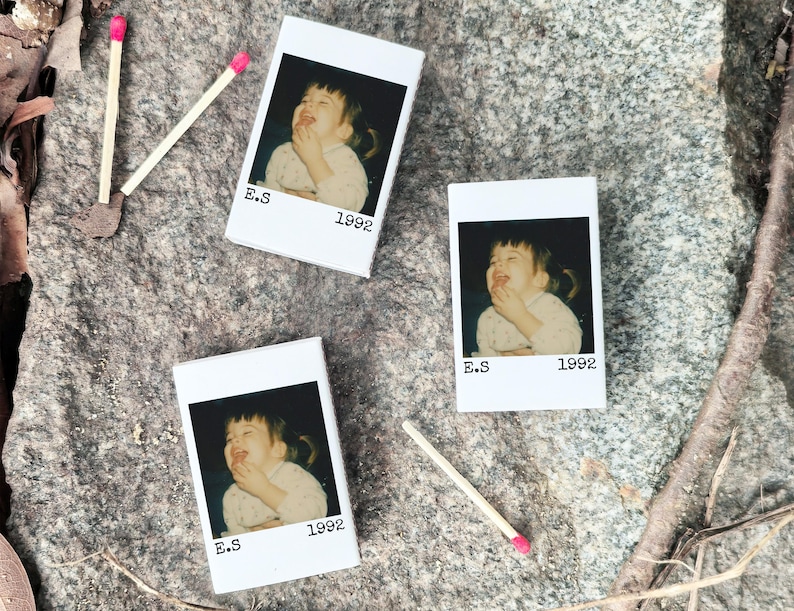May include: Three white matchbox-style containers with a photo of a laughing child, the text "E.S 1992". Two matches with pink tips are next to the containers. The items are on a grey stone surface.