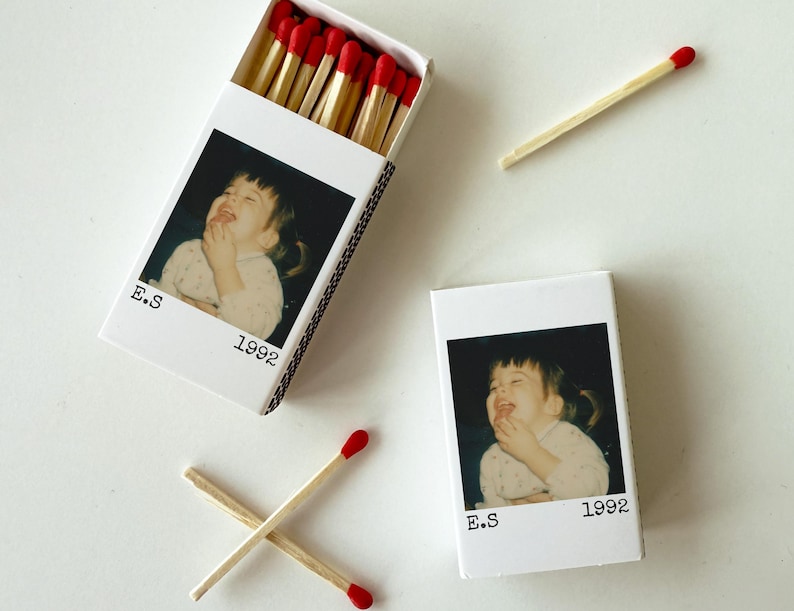 May include: Matchboxes featuring a photograph of a laughing child, labelled "E.S 1992". The boxes are white, with several matches visible, some inside the open box and others scattered nearby, with red tips.