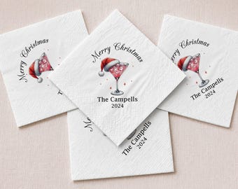 May include: White square paper napkins featuring a printed illustration of a martini glass filled with a pink drink and topped with a red Santa hat. The text reads Merry Christmas, The Campells, and 2024 in black lettering.