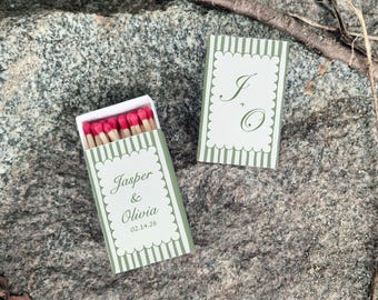 May include: Two matchboxes with green and white striped designs. One box is open, revealing red-tipped matches. The boxes feature the names "Jasper & Olivia" and initials "J + O", with a date. The boxes are on a gray stone surface.