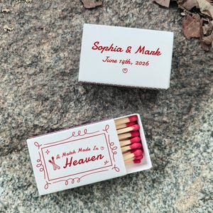 May include: Two white matchboxes with red text and accents. One box reads "Sophia & Mark June 14th, 2026" with a heart. The other box says "A Match Made In Heaven" with a decorative border and matches with red tips.