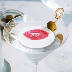 May include: A martini glass filled with a clear liquid and a white paper coaster with a red lipstick kiss print. A green olive is on a toothpick in the glass.