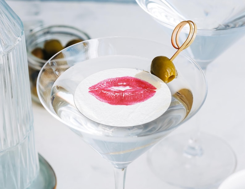 May include: A martini glass filled with a clear liquid and a white paper coaster with a red lipstick kiss print. A green olive is on a toothpick in the glass.