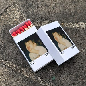 May include: Two white matchboxes, one open, displaying a photograph of a laughing child. The boxes are printed with "E.S" and "1992". The open box reveals red-tipped matches.