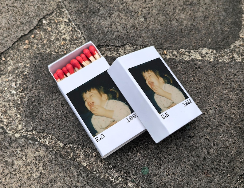 May include: Two white matchboxes, one open, displaying a photograph of a laughing child. The boxes are printed with "E.S" and "1992". The open box reveals red-tipped matches.