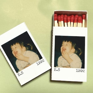 May include: A white matchbox and a Polaroid-style photo featuring a laughing child. The matchbox is open, revealing red-tipped matches. The photo and matchbox both have the text "E.S 1992".