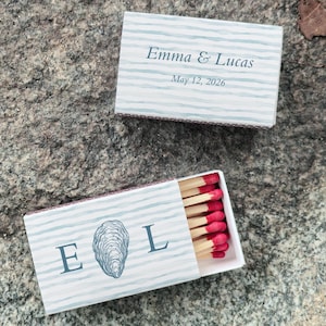 May include: Two matchboxes with a light blue and white striped design. One box is closed and has the text "Emma & Lucas May 12, 2026". The other box is open, revealing red-tipped matches, and features the letters "E L" with an oyster illustration.