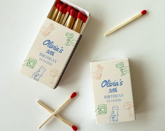 May include: Two matchboxes and loose matches. The matchboxes are off-white with blue text that reads "Olivia's 30th BIRTHDAY 03.14.2026". The boxes have colorful line drawings of cocktails. The matches have red tips.