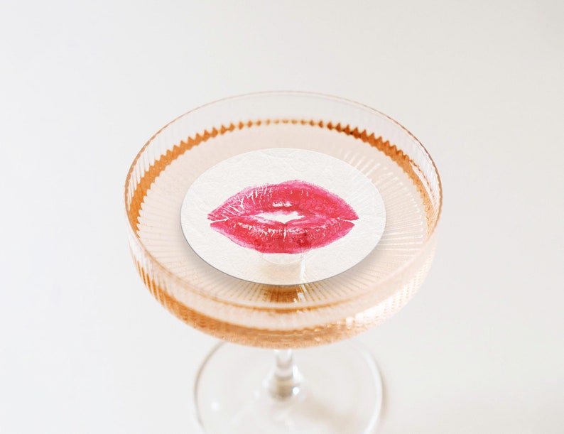 May include: A clear glass cocktail glass with a gold rim and a white paper coaster with a red lipstick kiss mark in the center.