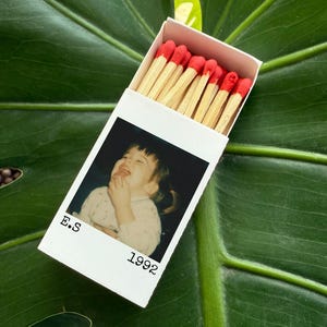 May include: A white matchbox with a photograph of a laughing child, the text "E.S 1992" is printed on the box. The match heads are red, and the matchsticks are natural wood colour. The matchbox rests on a large green leaf.