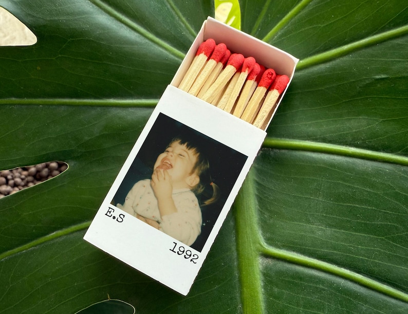 May include: A white matchbox with a photograph of a laughing child, the text "E.S 1992" is printed on the box. The match heads are red, and the matchsticks are natural wood colour. The matchbox rests on a large green leaf.