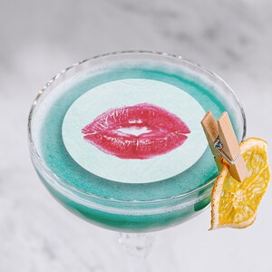 May include: A blue cocktail with a white paper circle with a red lipstick kiss print on top. A dried orange slice is attached to the glass with a wooden clothespin.
