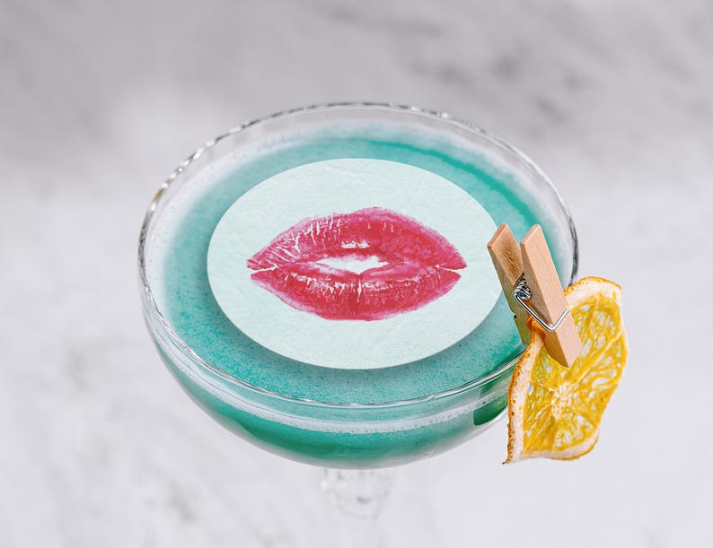 May include: A blue cocktail with a white paper circle with a red lipstick kiss print on top. A dried orange slice is attached to the glass with a wooden clothespin.