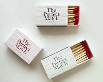 May include: Three matchboxes with the text "The Perfect Match" and the date 12.25.26. Two boxes are open, revealing red-tipped matches. The boxes have a white background with a striped design, and the text is in different colors.