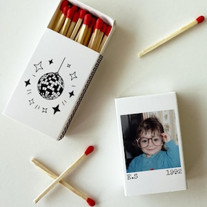 May include: Two white matchboxes with red-tipped matches. One box features a black line drawing of a disco ball surrounded by stars. The other box displays a color photograph of a child wearing glasses, with the text E.S and 1992 printed below.