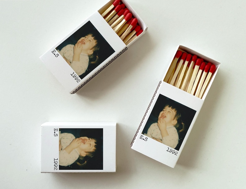 May include: Three white matchboxes, each featuring a photograph of a child. The boxes contain wooden matches with red tips. The text "E.S" and "1992" are printed on the front of each box, adding a personal touch.