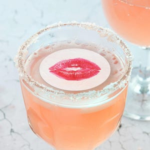 May include: A pink cocktail with a sugared rim and a white paper circle with a red lipstick kiss print on top.