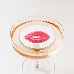 May include: A clear glass champagne flute with a white paper coaster featuring a red lipstick kiss mark.