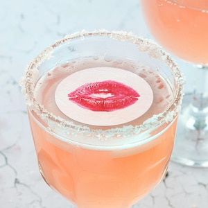 May include: A pink cocktail with a sugared rim and a white paper circle with a red lipstick kiss print on top.