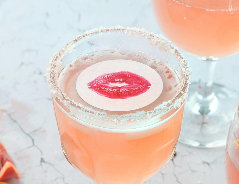 May include: A pink cocktail with a sugared rim and a white paper circle with a red lipstick kiss print on top.