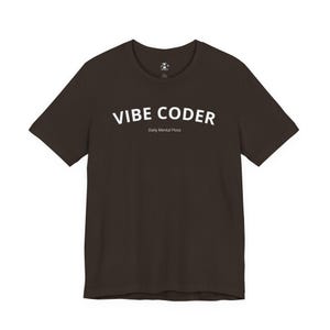 Op de afbeelding: Een donkerbruin t-shirt met de witte tekst "VIBE CODER" en "Daily Mental Floss" op de voorkant. Het shirt heeft korte mouwen en een ronde hals.
