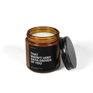 May include: A brown glass candle jar with a black label that reads "That wasn't very data-driven of you". The candle is lit and the lid is off.