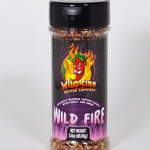 May include: A glass jar with a black lid filled with red pepper flakes. The label is purple with a red chili pepper and the text "WILD SIDE PEPPER COMPANY" and "WILD FIRE". The net weight is 3.0oz (85.05g).
