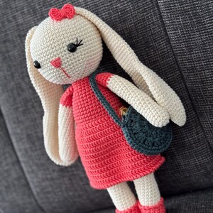 Peut inclure: Une poupée de lapin en crochet portant une robe rouge et un sac à main turquoise. Le lapin a un nœud rose sur la tête et des yeux noirs.