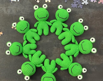 Op de afbeelding: Een verzameling van acht handgemaakte groene gehaakte kikker speelgoed, gerangschikt in een cirkel. Elke kikker heeft witte en zwarte ogen en een lachende mond. De achtergrond bevat verspreide puzzelstukjes.