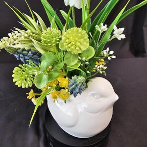 May include: A white ceramic bird-shaped vase filled with an arrangement of artificial flowers. The flowers include white tulips, yellow, green, and blue blossoms, and various green foliage. The vase sits on a black base.