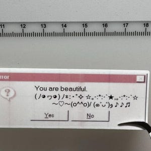Op de afbeelding: Een witte sticker met een roze bovenrand, die lijkt op een foutmelding. De tekst luidt "You are beautiful" met verschillende symbolen en de opties "Yes" en "No". Een liniaal staat op de achtergrond.