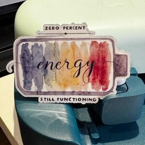 May include: A battery-shaped sticker with watercolour stripes in blue, yellow, and red. The word "energy" is written in cursive, and the text "ZERO PERCENT" and "STILL FUNCTIONING" are also included.