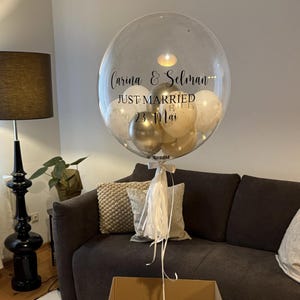 Könnte beinhalten: Ein transparenter Blasenballon mit goldenen und weißen Ballons im Inneren. Der Ballon trägt eine schwarze Aufschrift mit dem Text "Carina & Selman Just Married 23 Mai". Der Ballon befindet sich in einem braunen Karton mit der Aufschrift "Bubble Fabrik" auf der Seite.