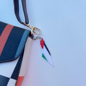 May include: A purse with a striped pattern in red, black, and blue, featuring a black strap and gold-coloured clasp. A charm in the shape of the Palestinian flag, with red, white, green, and black, is attached.