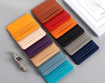 May include: A collection of colorful, slim card wallets in various shades including orange, brown, red, black, gray, and blue. Each wallet has multiple card slots. A silver wristwatch is also visible.