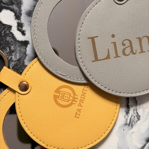 以下が含まれることがあります： 3つの丸いコンパクトミラーのクローズアップ。1つは黄色、1つは「Lian」という名前がプリントされたグレー、もう1つはグレーです。それぞれに金属製のリングと小さな鏡が付いています。